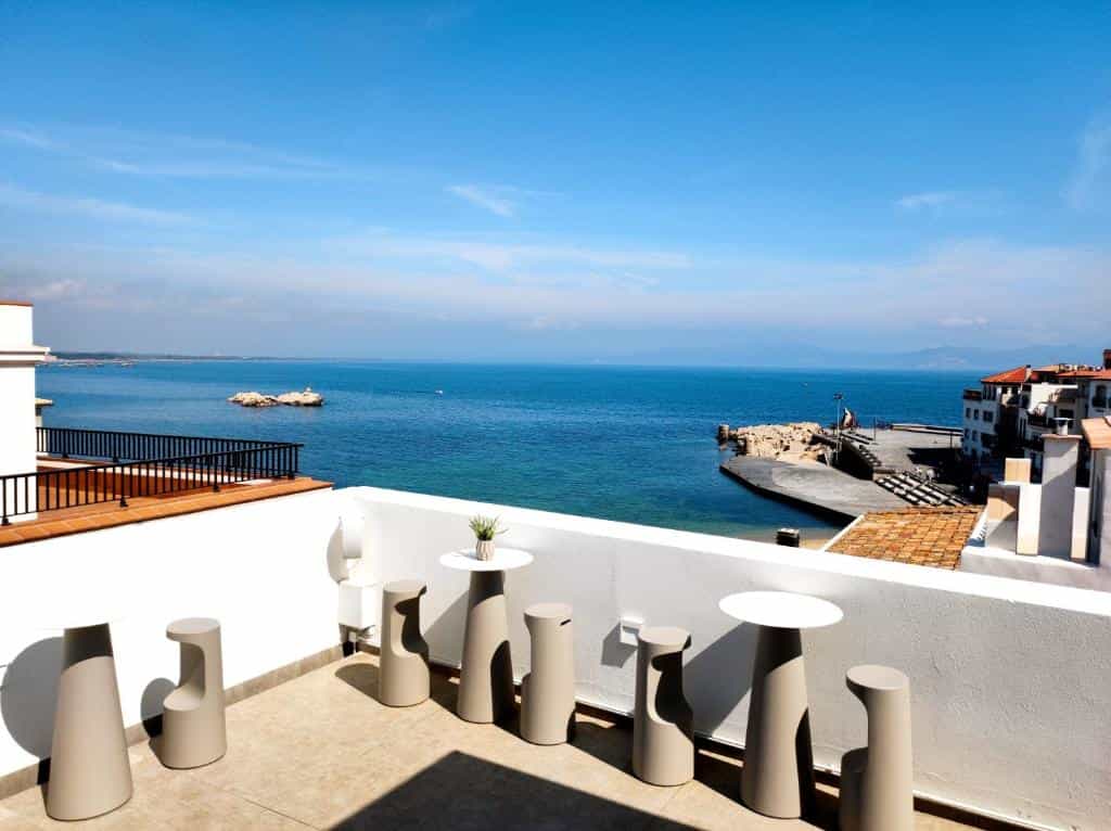Rooftop terrace with modern tables and stools overlooking a coastal town and the Mediterranean Sea.
