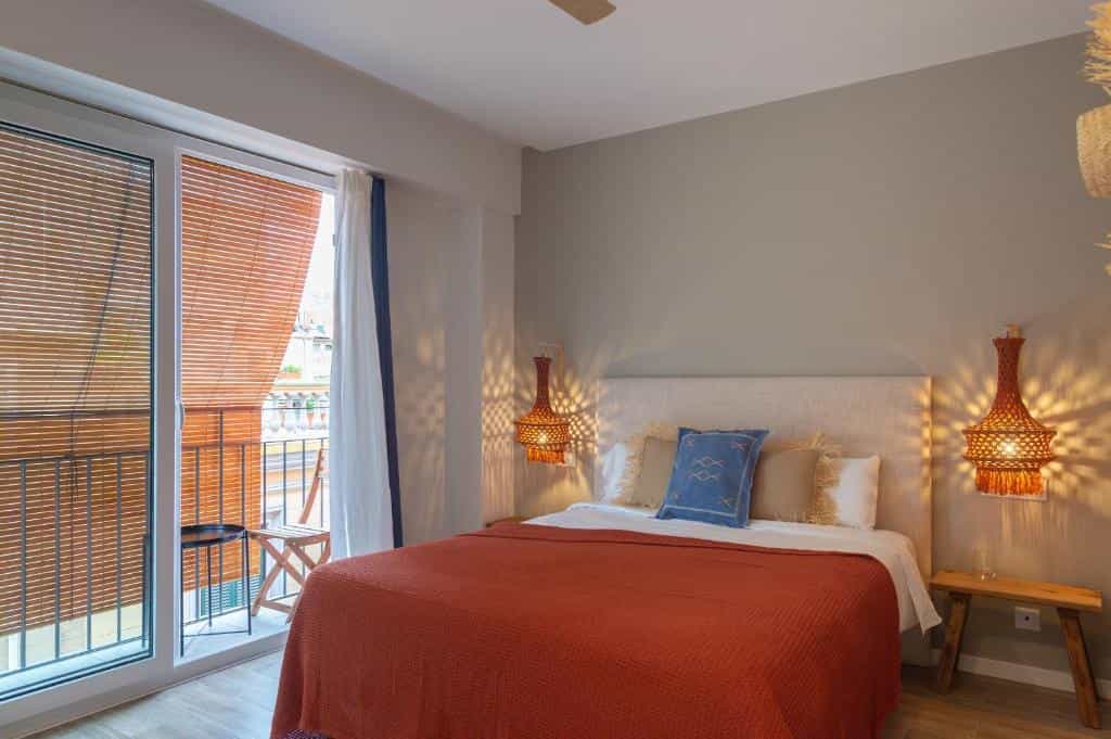 Hotel bedroom with a double bed, woven pendant lights, and a balcony overlooking a city street.