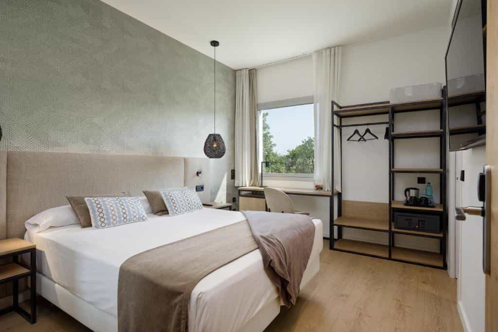Modern hotel room with a double bed, patterned wallpaper, open wardrobe, and a desk by the window.