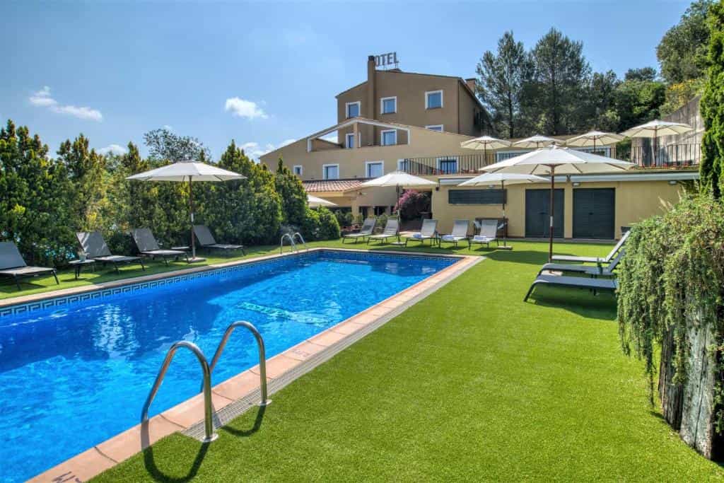 Swimming pool with sun loungers and umbrellas on a green lawn in front of a hotel building.