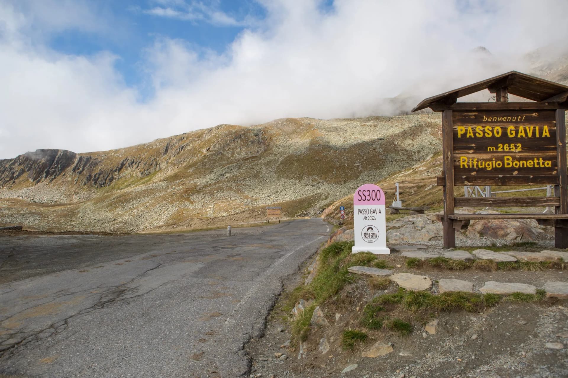 Passo Gavia m. 2652 sign next to cracked mountain road with rocky slopes and clouds.
