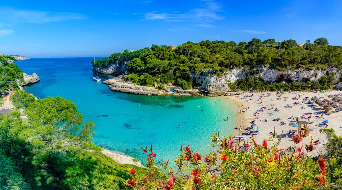 Cyclists on a coastal road overlooking a turquoise cove with a busy beach and sailboat.