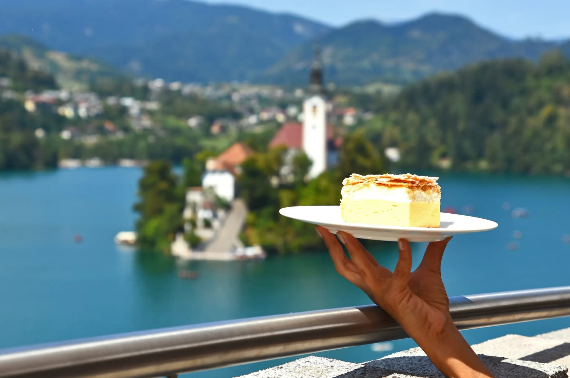 Cream cake slice held on a plate overlooking Lake Bled island church and mountains in Slovenia.