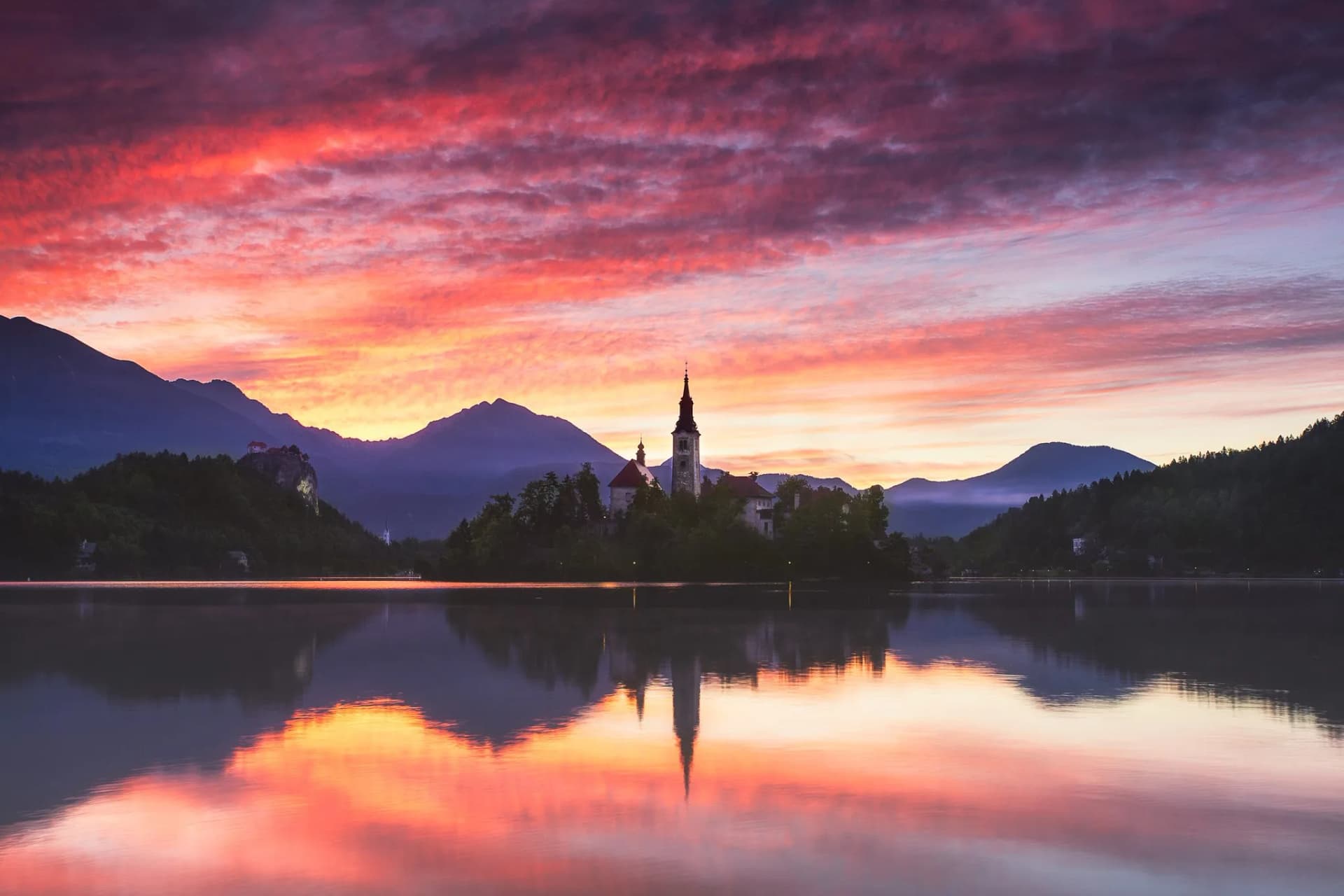 Lake Bled island church reflected in water at sunrise with mountains and pink sky