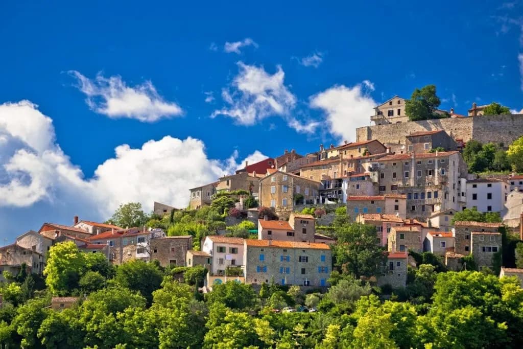 Hilltop village with stone houses and red roofs nestled in green trees under a bright blue sky.