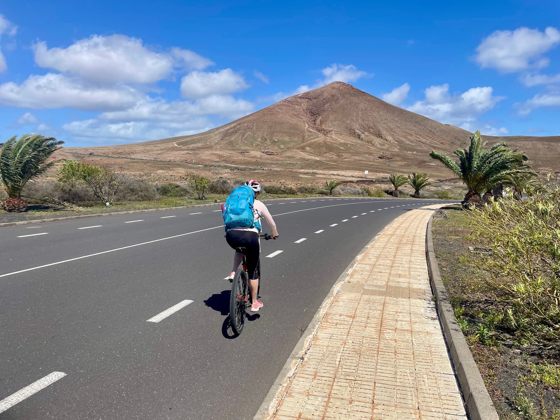Cyclist with a blue backpack riding on a paved road toward a volcanic mountain in Lanzarote.