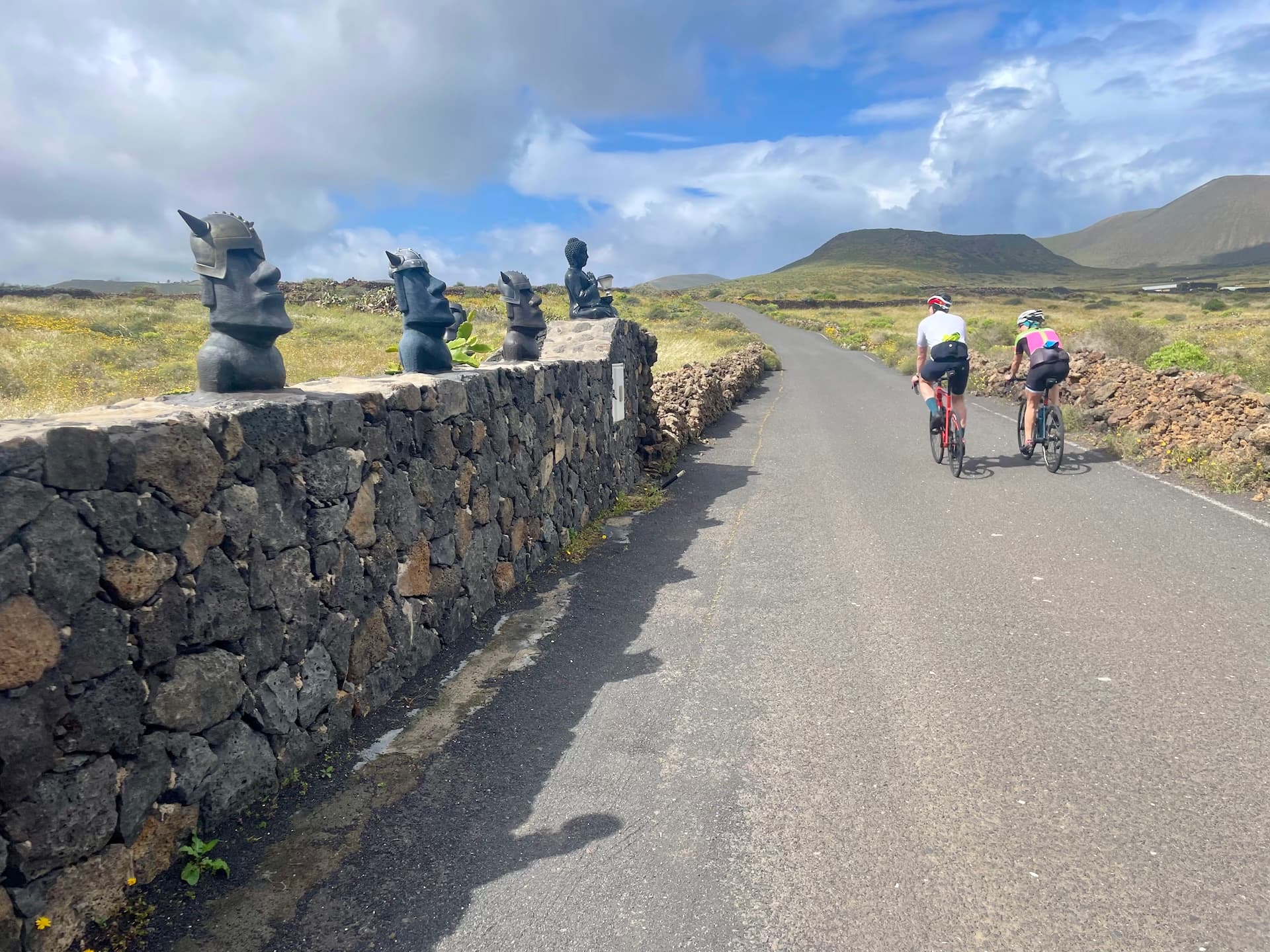 Two cyclists riding on a road in Lanzarote past a stone wall topped with decorative statues and volcanic landscape.