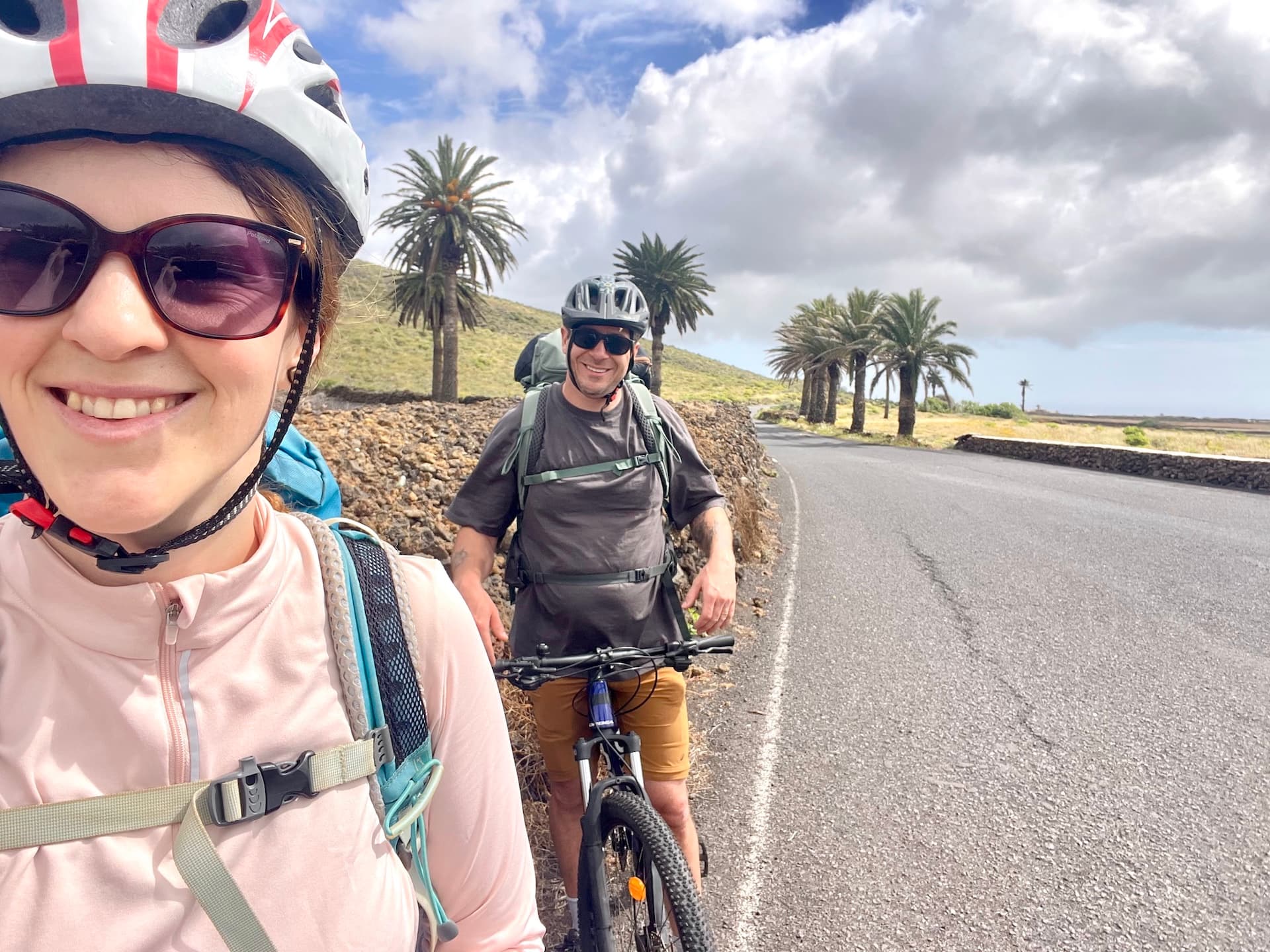 Two cyclists with backpacks on a road lined with palm trees in Lanzarote.