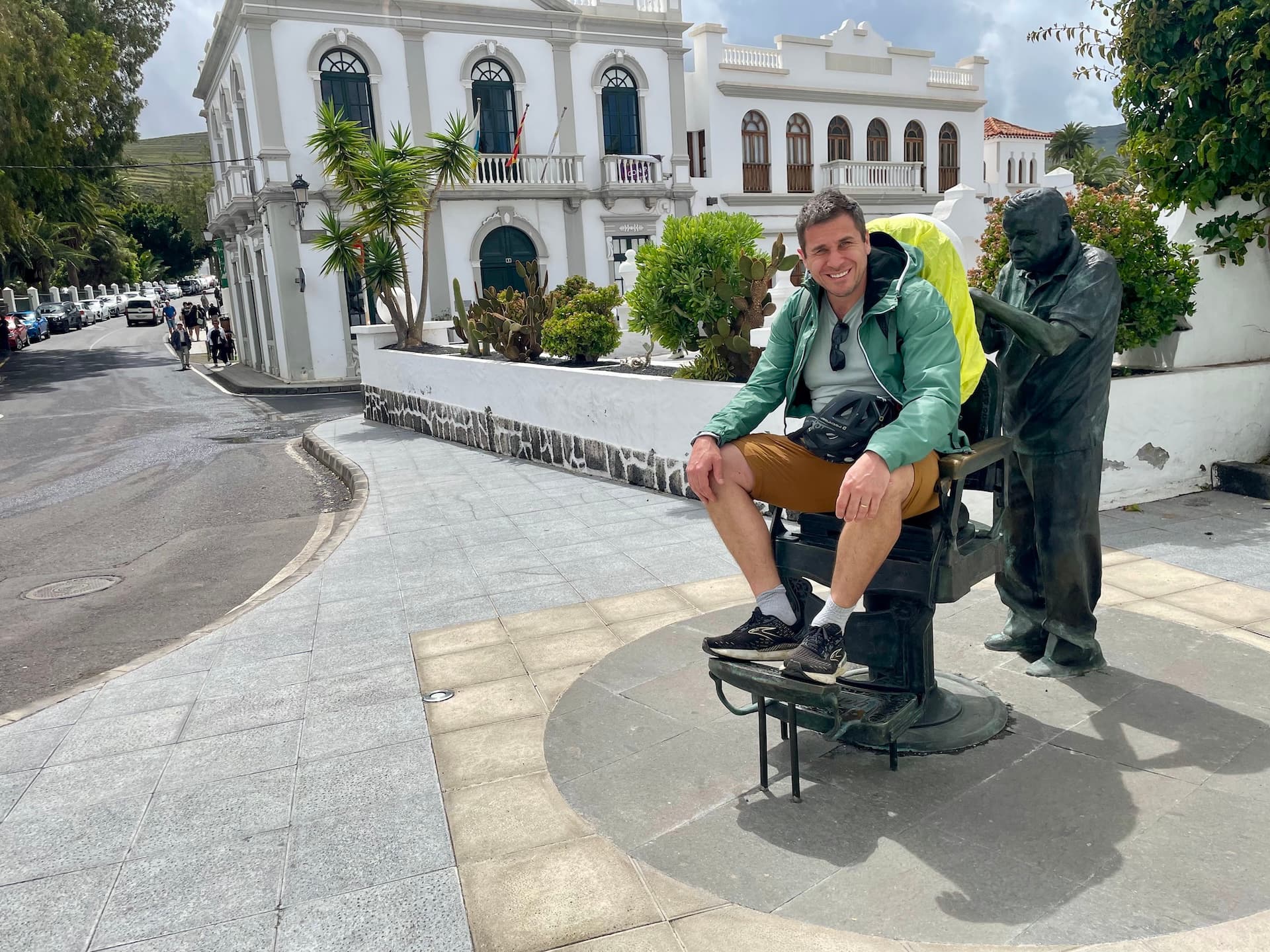 Cyclist sitting in a barber chair statue in Haría, Lanzarote, next to a white colonial-style building.