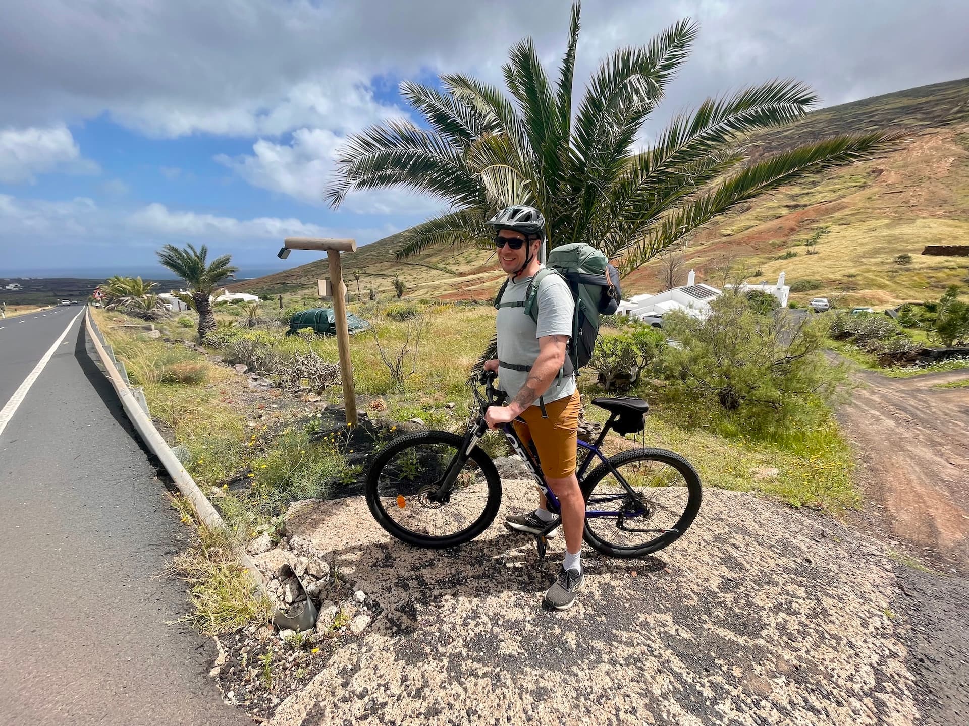Cyclist with backpack standing on a roadside in Lanzarote with palm trees and volcanic hills.