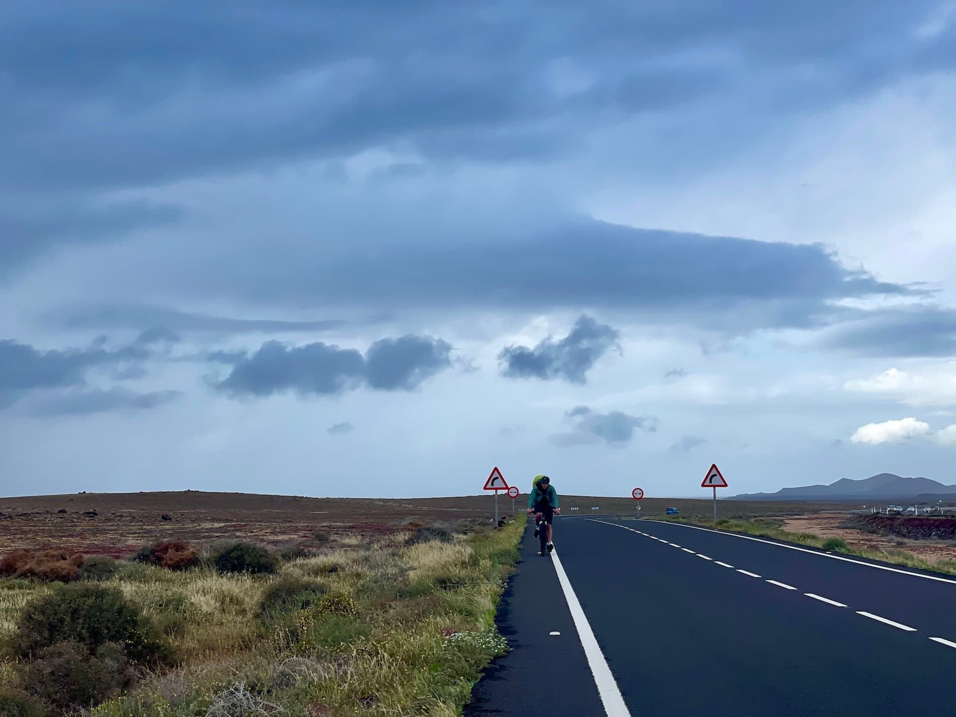 Cyclist riding on an open road through a volcanic landscape in Lanzarote under a cloudy sky.