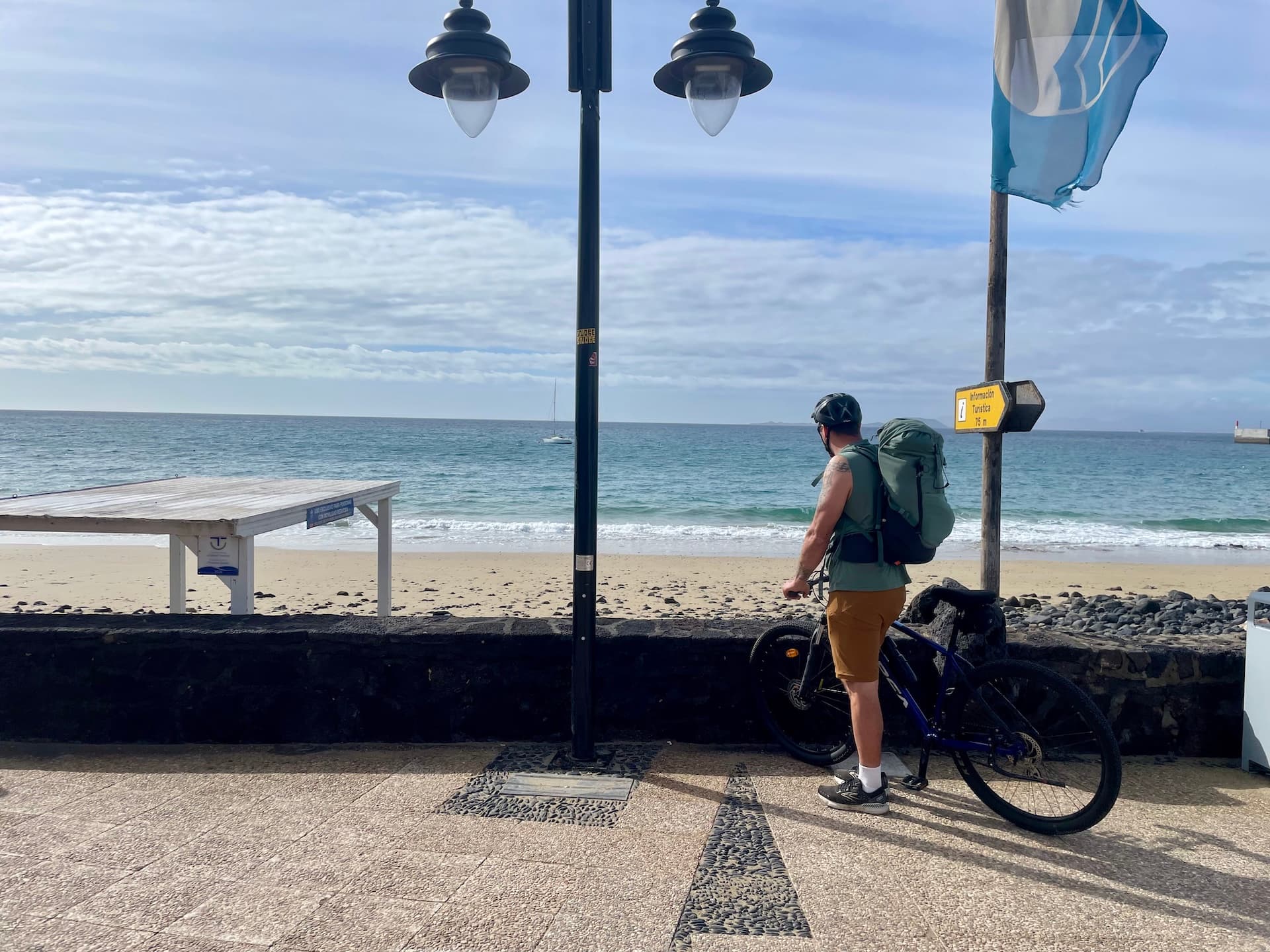 Cyclist with a backpack standing by a bicycle on a promenade overlooking the beach in Lanzarote.