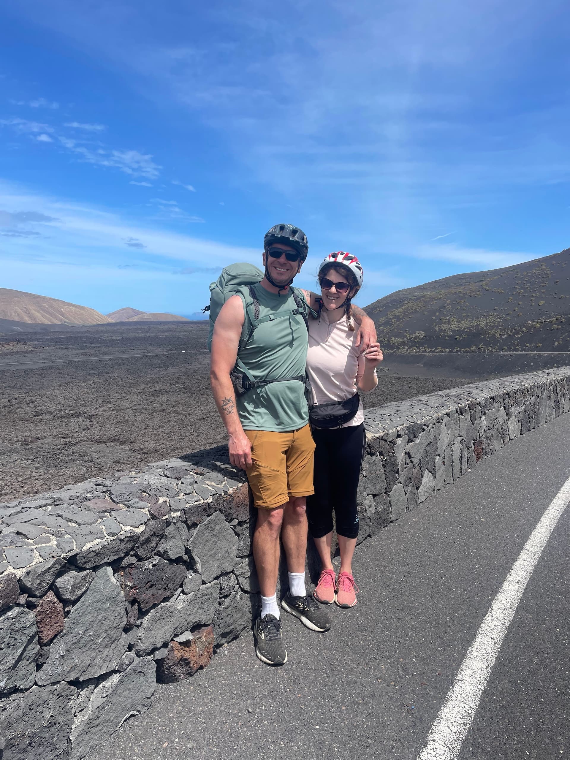 Two cyclists wearing helmets standing by a stone wall in a volcanic landscape in Lanzarote.