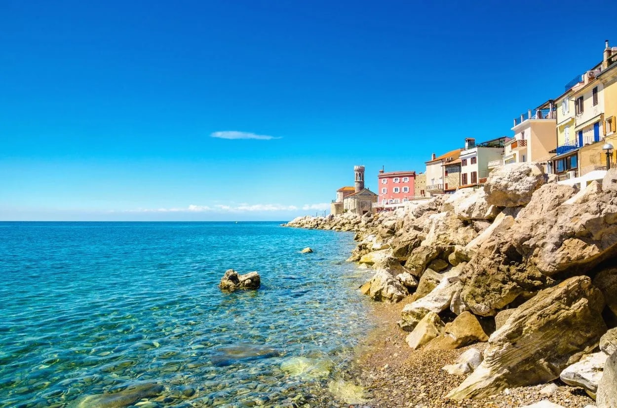 Piran coastline with colorful buildings and clear turquoise water under a bright blue sky.