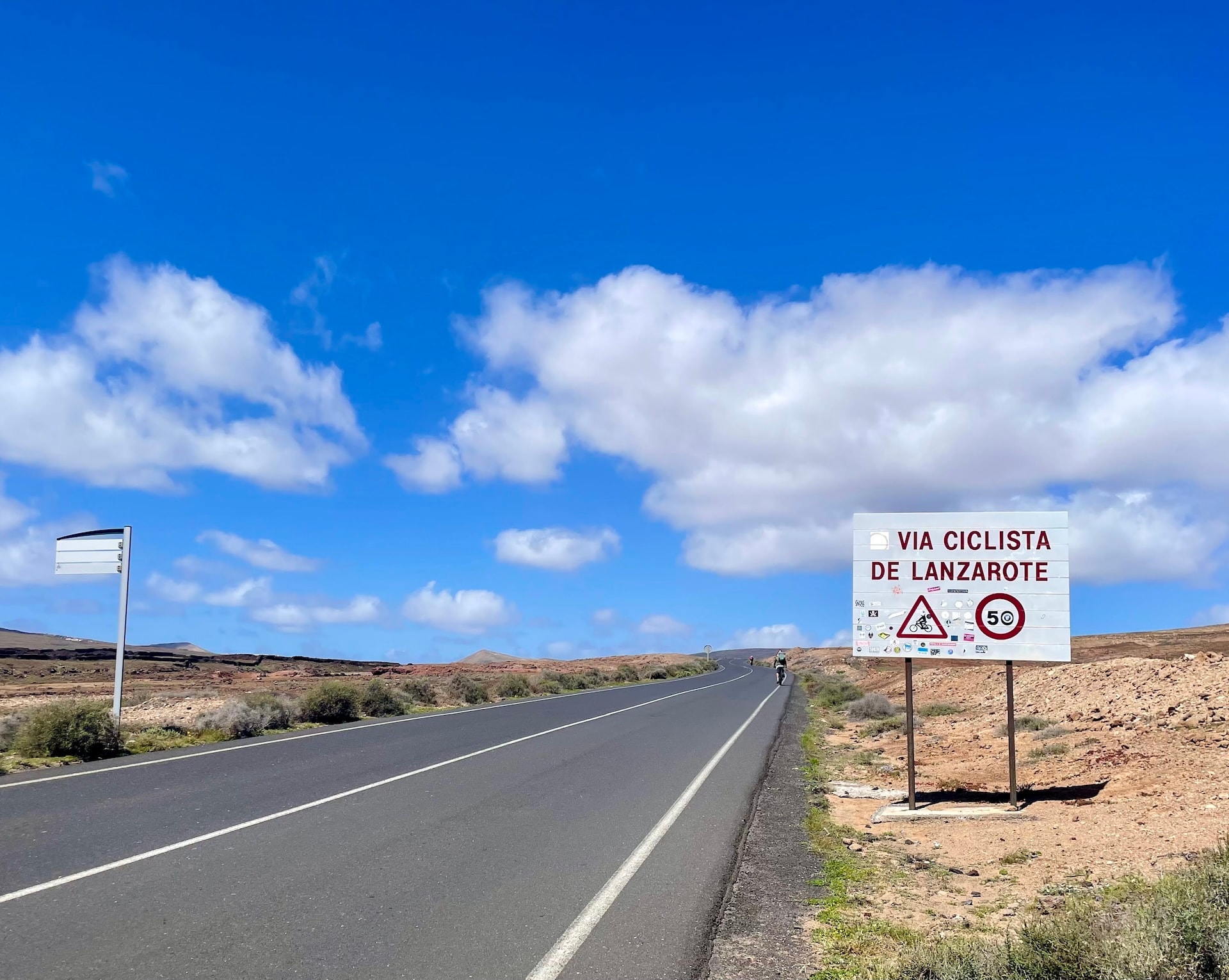 Cyclist on a paved road in Lanzarote with a Via Ciclista de Lanzarote sign under a blue sky.
