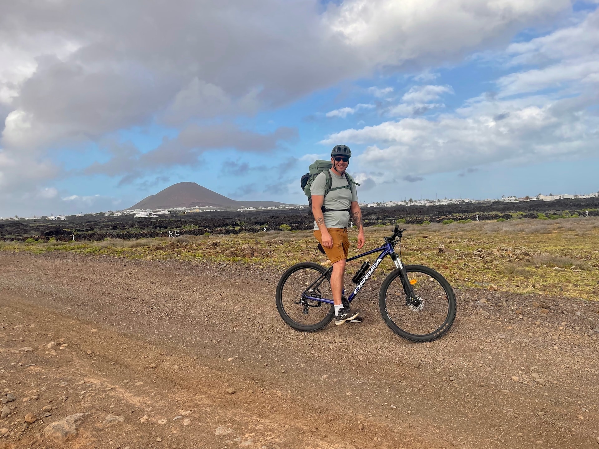 Cyclist with backpack on a gravel path in Lanzarote with volcanic landscape and a mountain in the background.