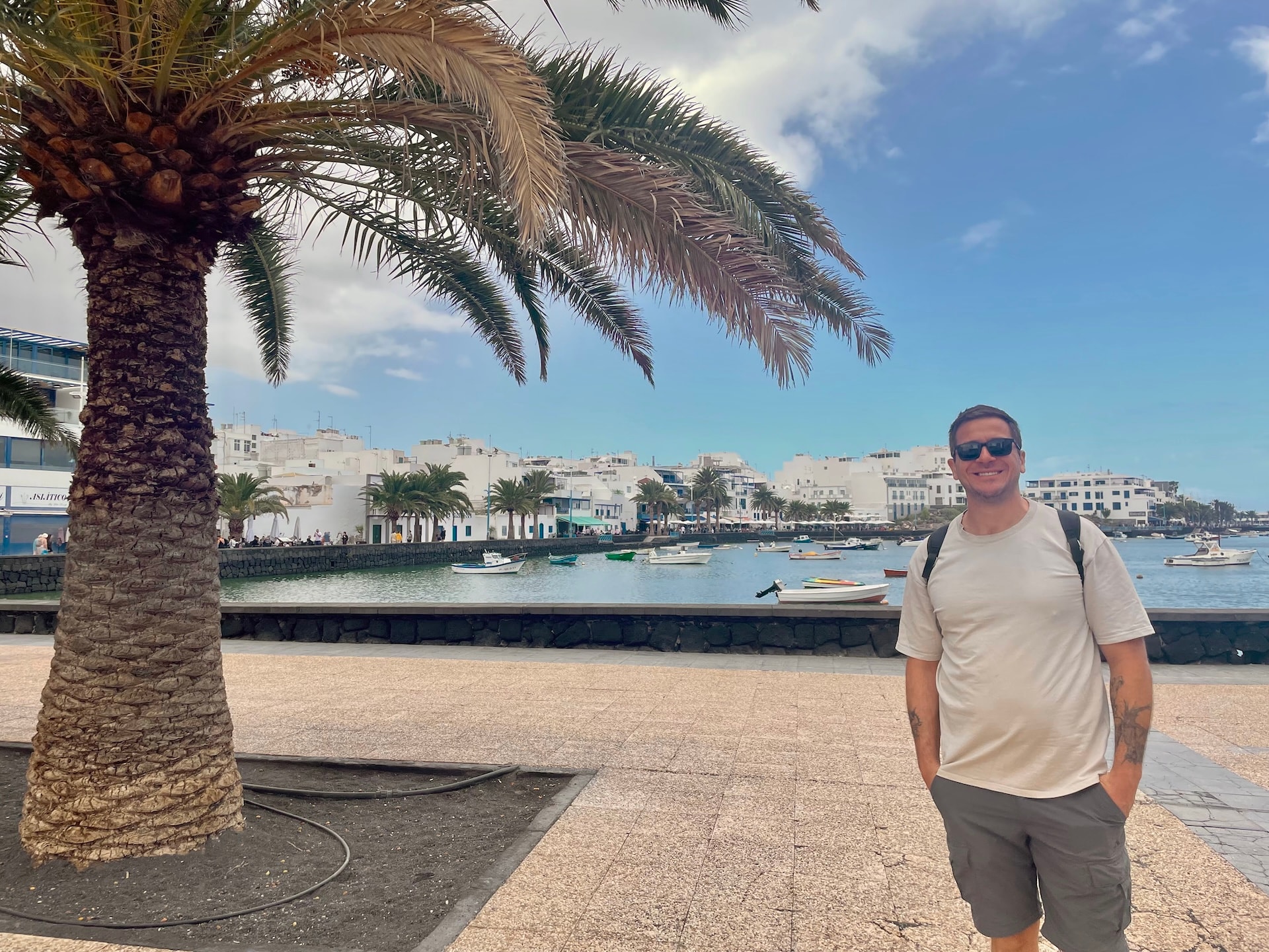 Man standing on a promenade by the harbor in Lanzarote with palm trees, white buildings, and small boats on the water.