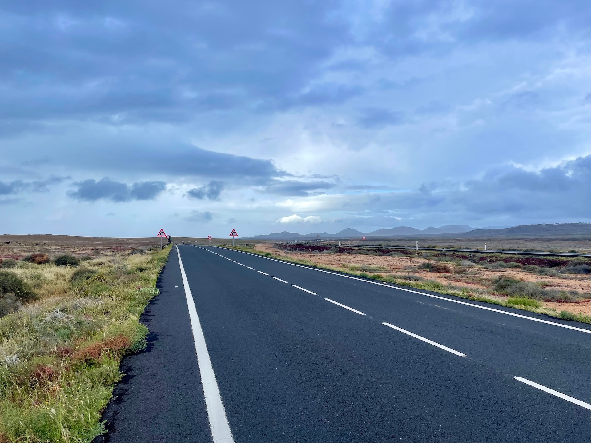 Empty asphalt road stretching through a volcanic landscape in Lanzarote under a cloudy sky.