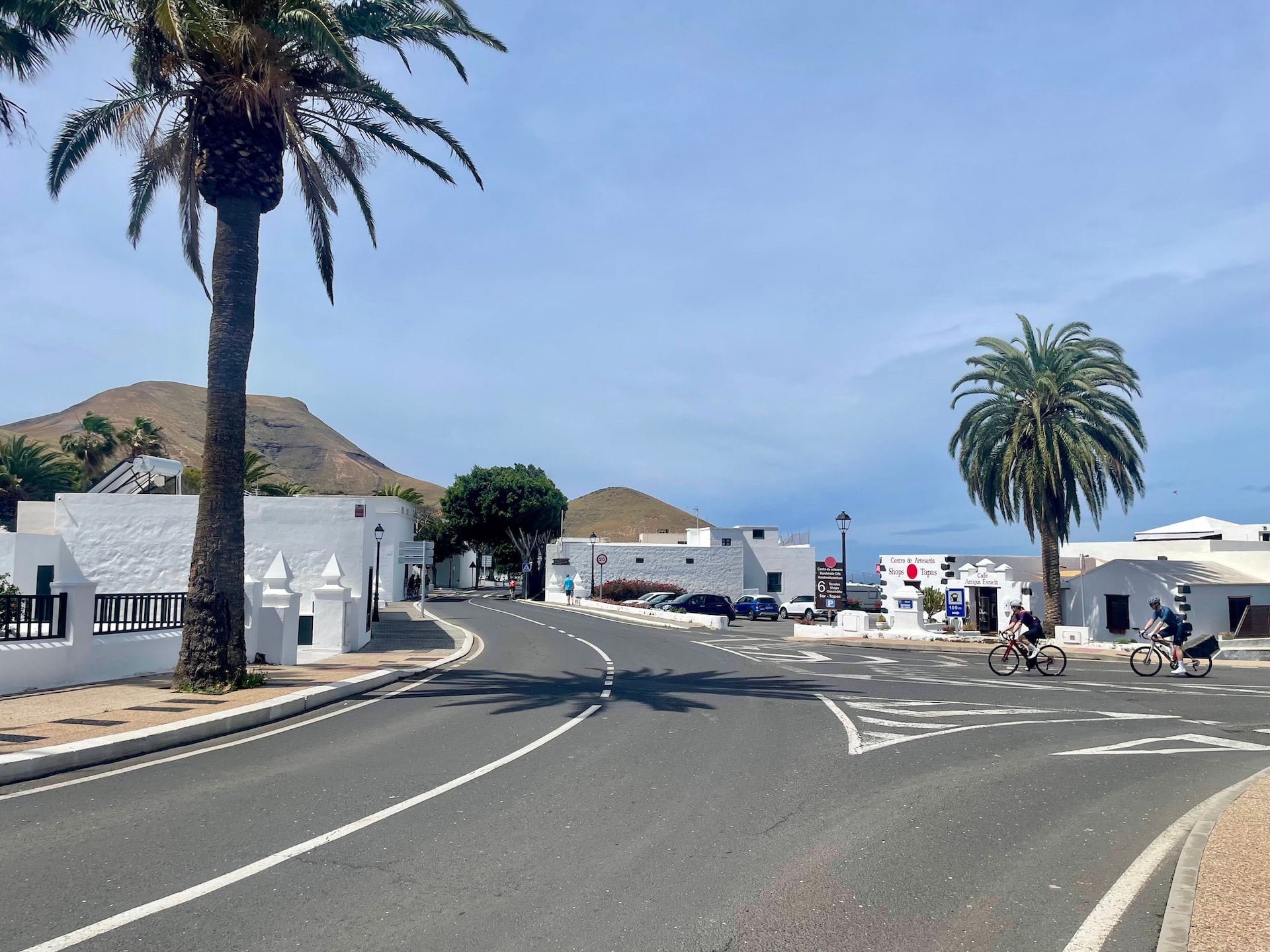 Cyclists riding on a road in Lanzarote with white buildings, palm trees, and volcanic hills in the background.