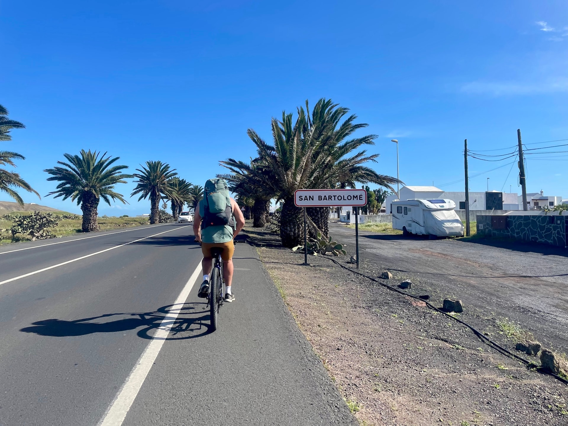 Cyclist with a backpack riding on a road past a San Bartolomé town sign lined with palm trees.
