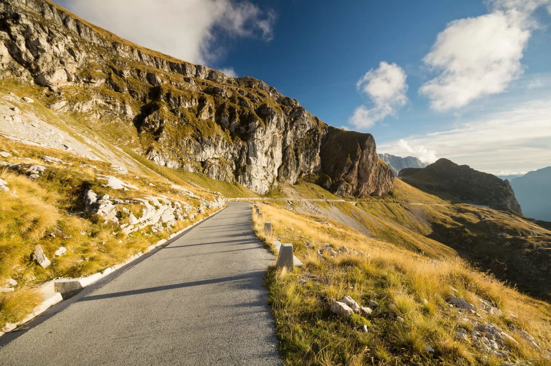 Mountain road winding past steep cliffs with dry yellow grass under a blue sky in the Julian Alps.