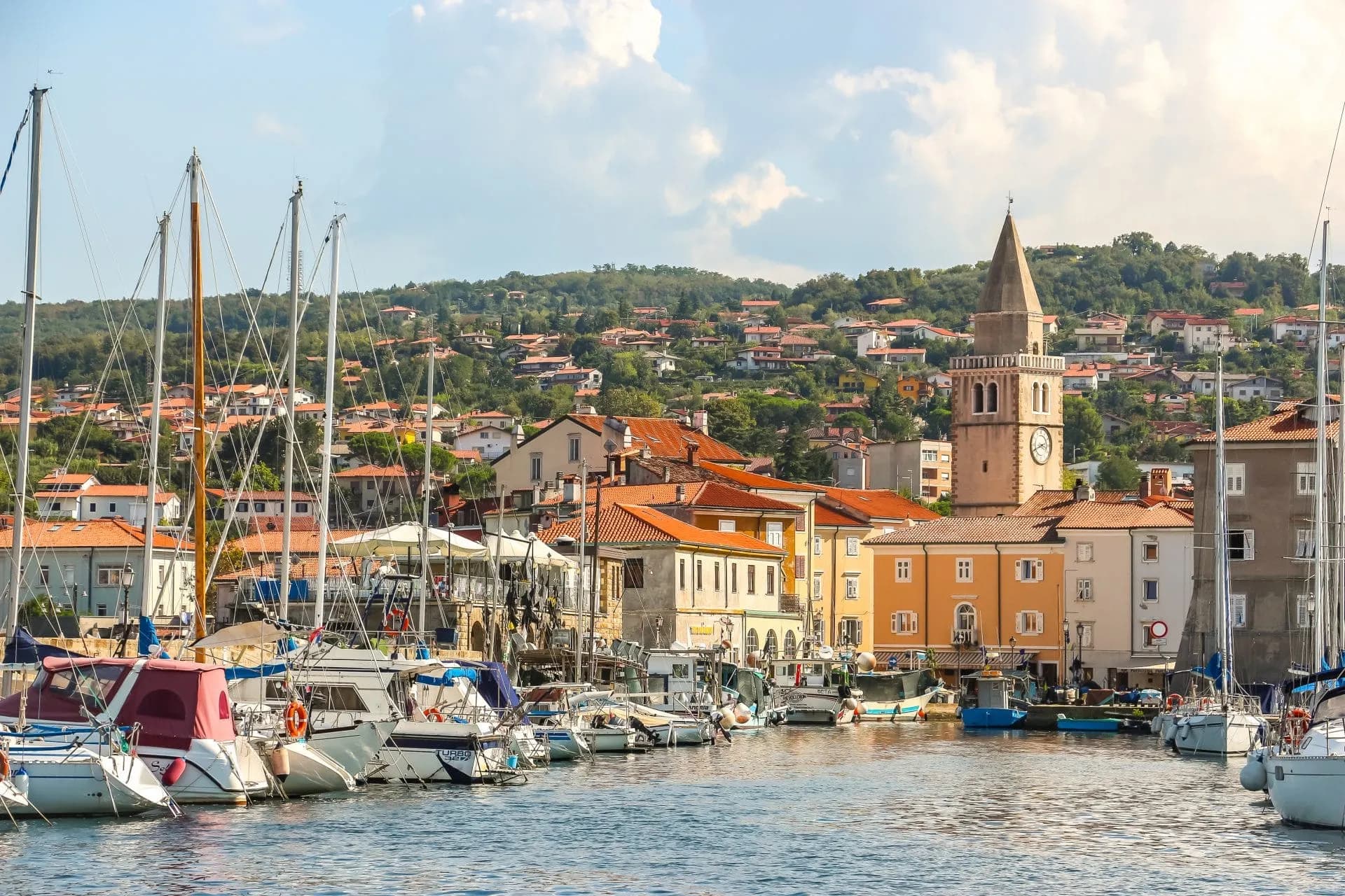 Sailboats docked in the harbor of Muggia, Italy, with colorful buildings and a clock tower backdrop.