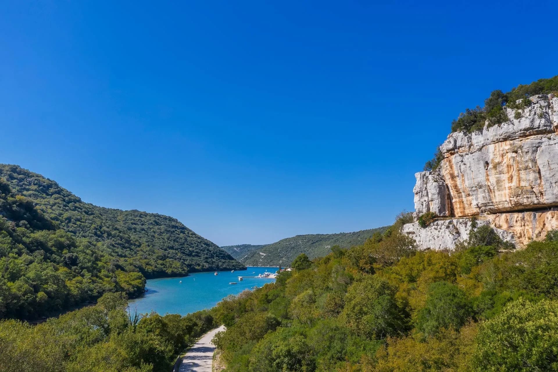 Turquoise inlet surrounded by lush green hills and a sheer limestone cliff under a clear blue sky.