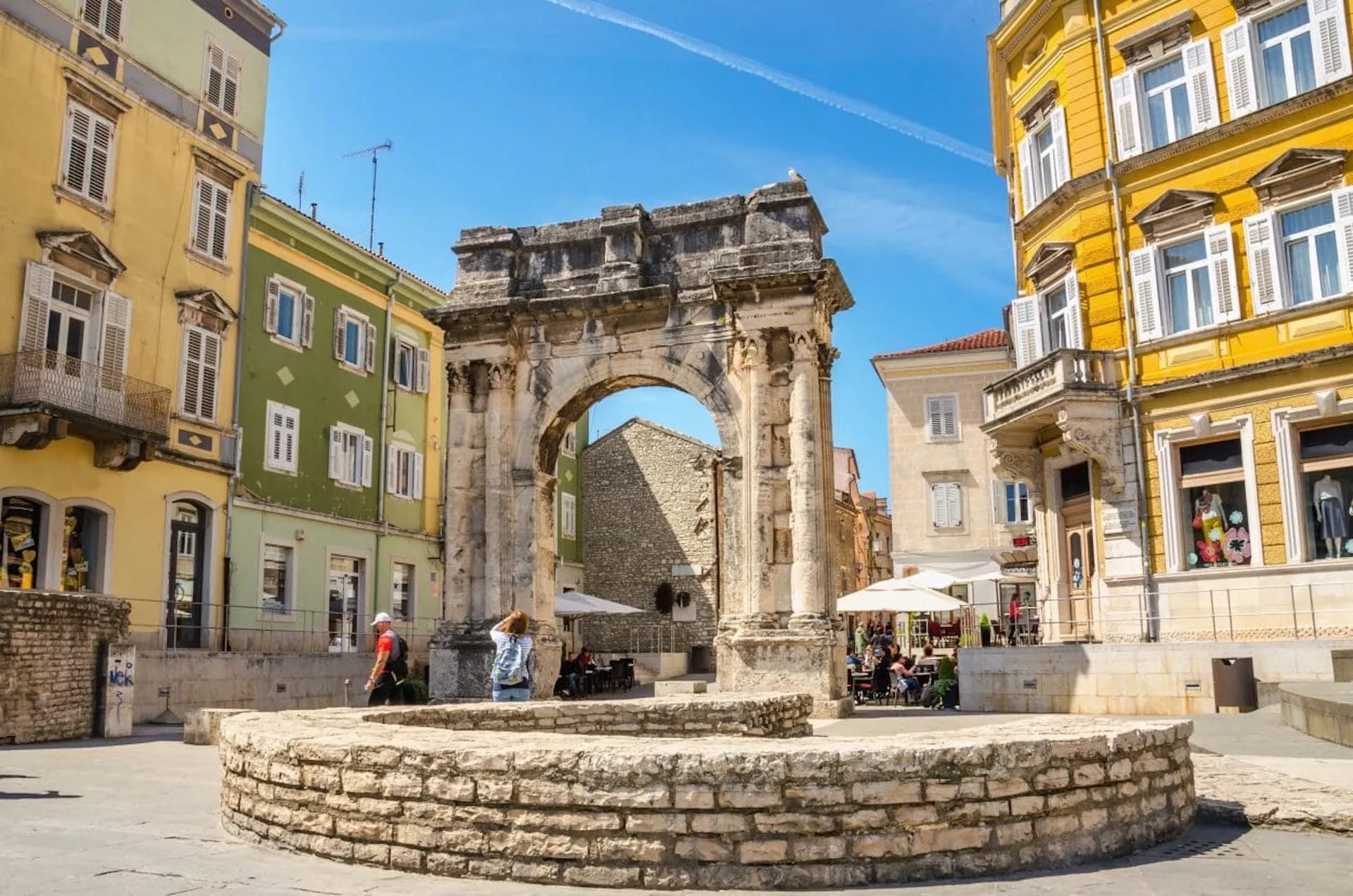 Triumphal arch in a square with colorful buildings and outdoor cafe seating in Pula.