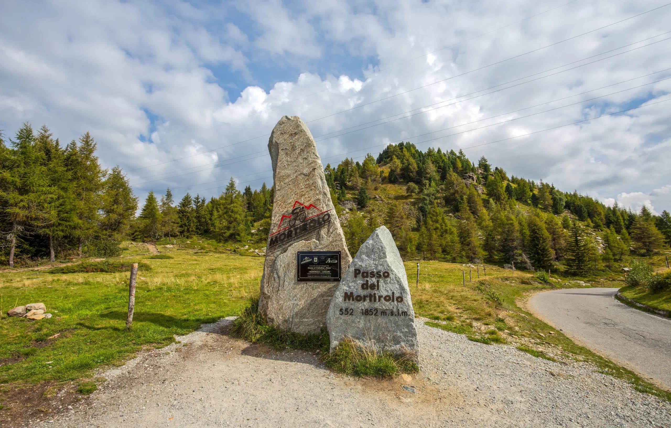 Passo del Mortirolo monument marker with green hillside, pine trees, and winding mountain road.