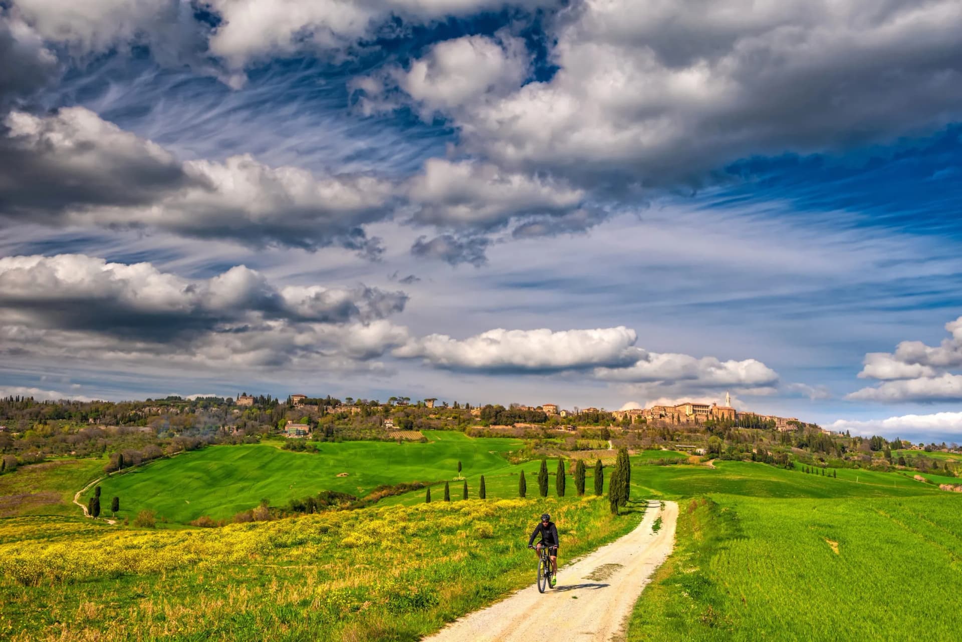 Cyclist on dirt road through rolling green hills toward Tuscan hilltop town under dramatic clouds.