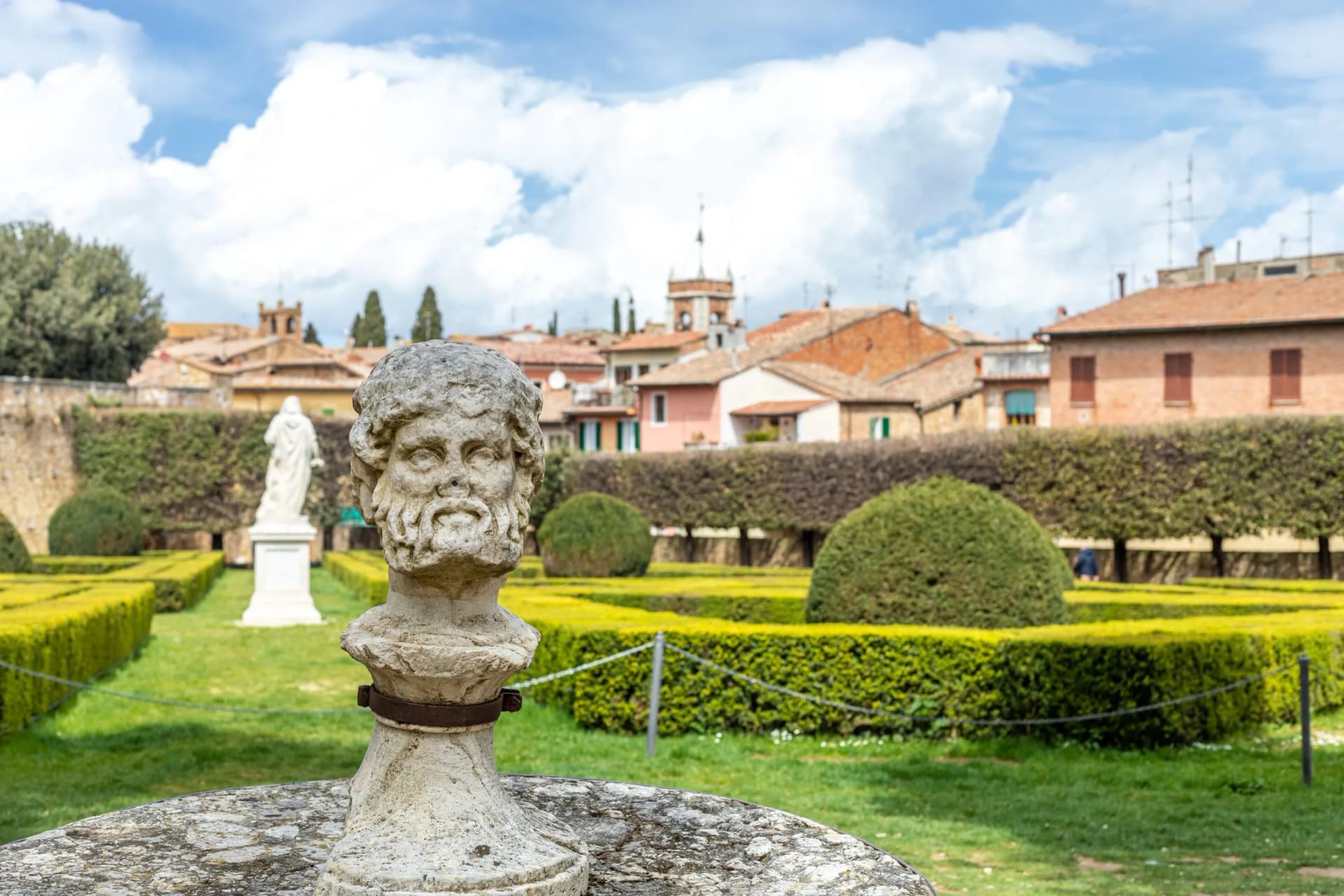 Stone bust in formal garden with manicured hedges and town buildings in San Quirico d'Orcia.