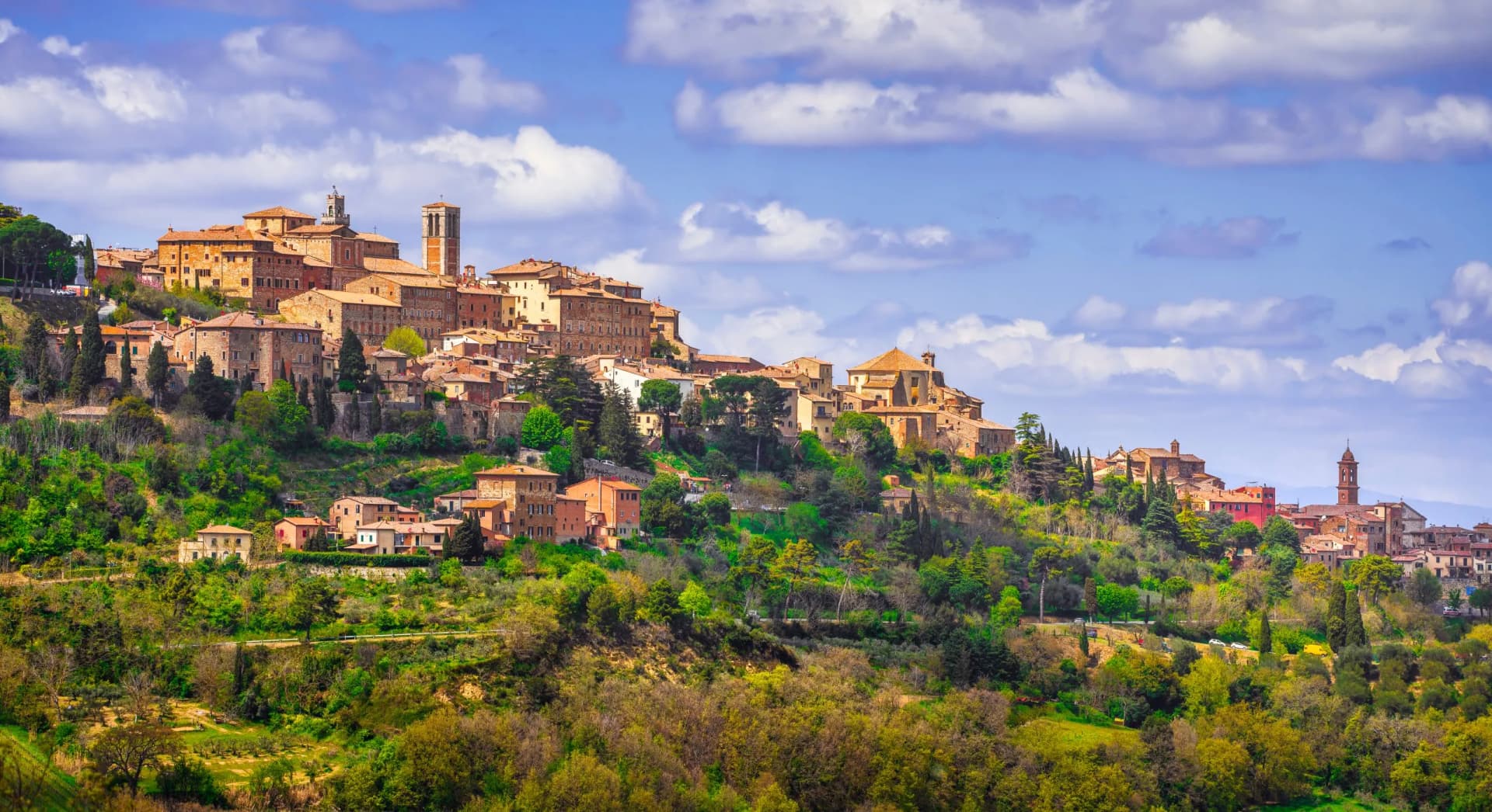 Hilltop Italian town with terracotta roofs nestled in lush green spring landscape under blue sky.