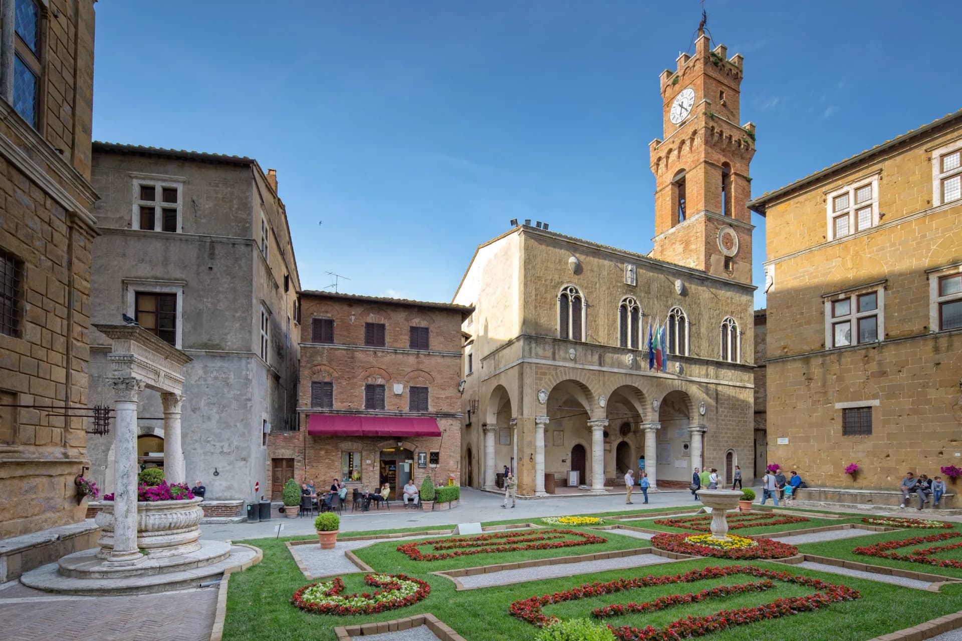 Pienza town square with historic buildings, clock tower, and manicured flower gardens under blue sky.