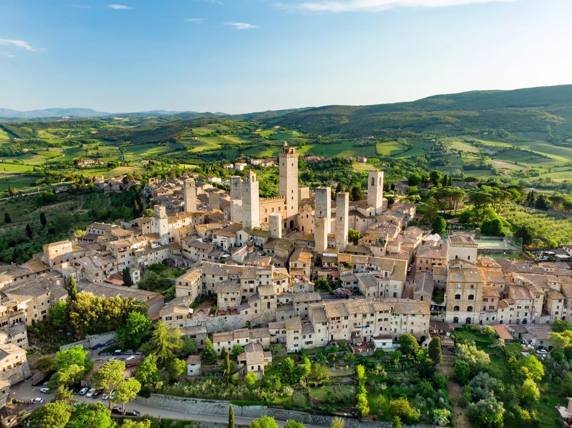 Aerial view of San Gimignano medieval town with stone towers surrounded by green Tuscan hills under a blue sky.