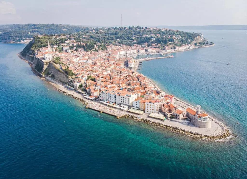 Aerial view of Slovenian coast and Piran peninsula with terracotta roofs meeting turquoise sea.