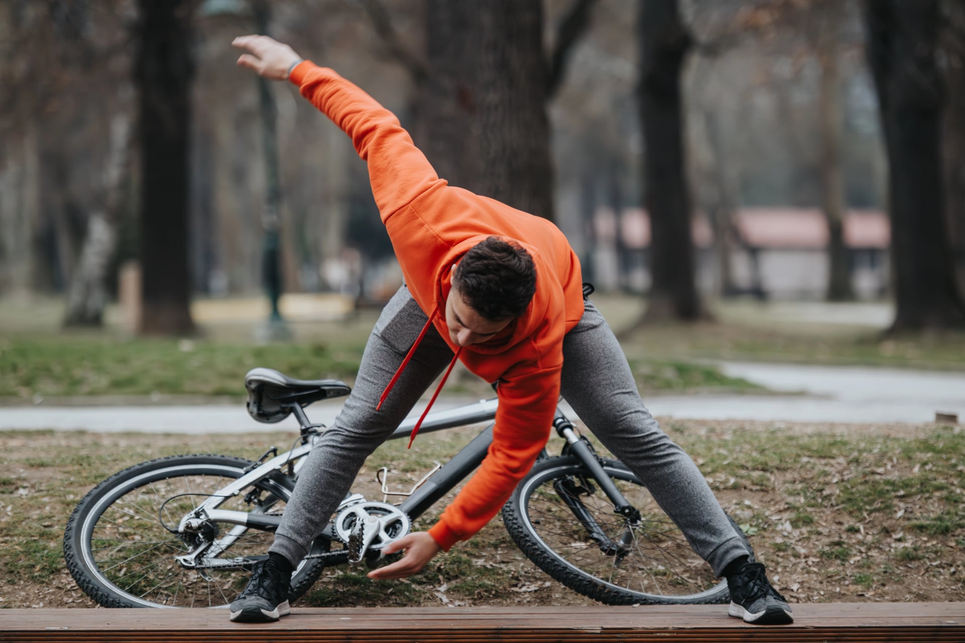 A man in athletic gear stretches alongside his bicycle in a park on a cloudy day, promoting health and fitness.