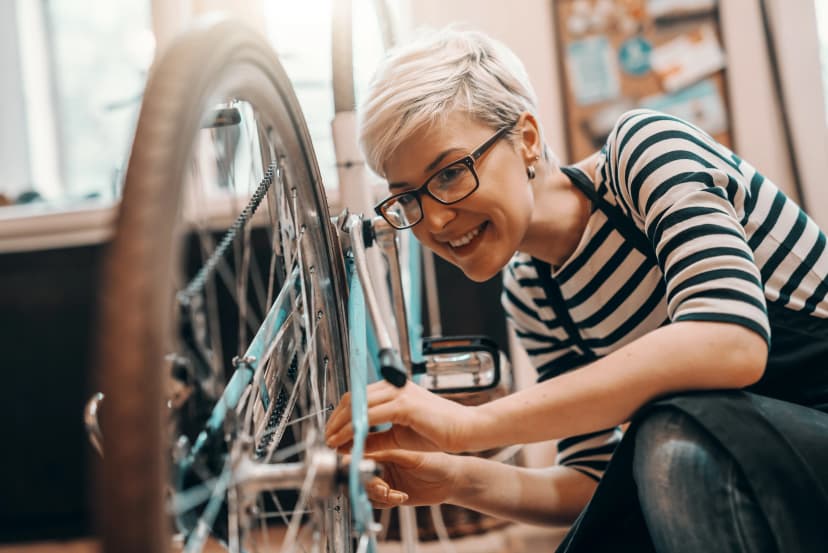 Beautiful Caucasian female worker with short blonde hair and eyeglasses crouching and repairing bicycle. Bike workshop interior.