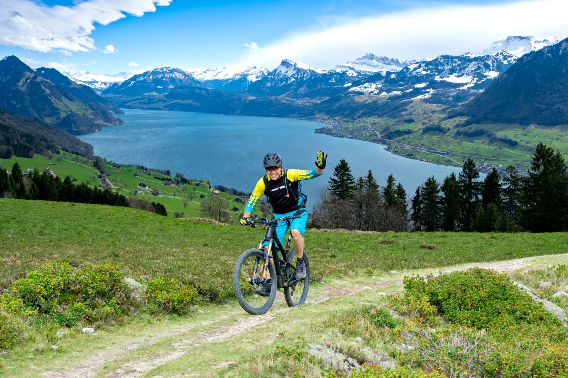 Sportive man in middle age with mountain bike on mountain trail beckons the viewer. The background shows the Lake Lucerne.